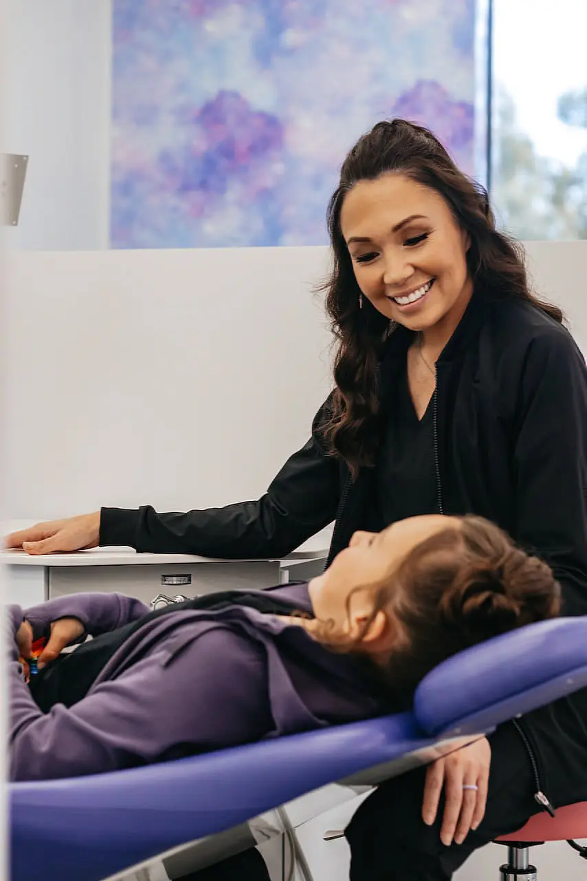 At Stellar Orthodontics in San Marcos CA, a woman in a black jacket smiles while discussing Phase I with a child reclining in a brightly lit dental chair.