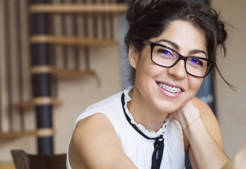 A woman with dark hair in buns, glasses, and a white sleeveless top smiles confidently with braces, while a spiral staircase is blurred in the background. Begin your orthodontic journey at Stellar Orthodontics in San Marcos, CA.