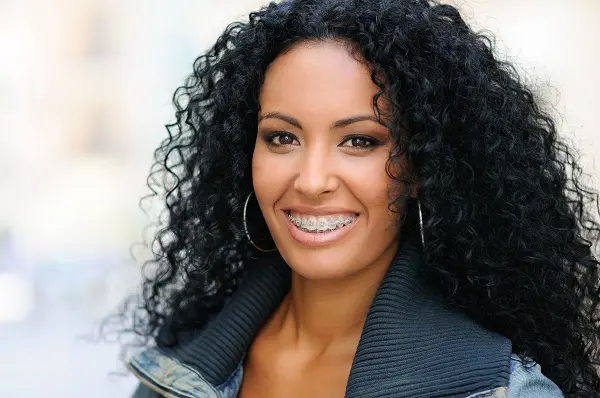 At Stellar Orthodontics in San Marcos CA, a woman with long curly black hair, hoop earrings, and a jacket smiles confidently, showing her metal braces and highlighting the advantages of adult orthodontics.