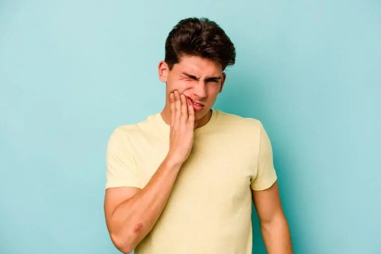 A young man in a yellow t-shirt stands against a blue background, wincing and holding his cheek—a reminder that urgent dental issues can arise unexpectedly. Stellar Orthodontics in San Marcos, CA offers emergency orthodontic care for sudden toothaches or facial discomfort.