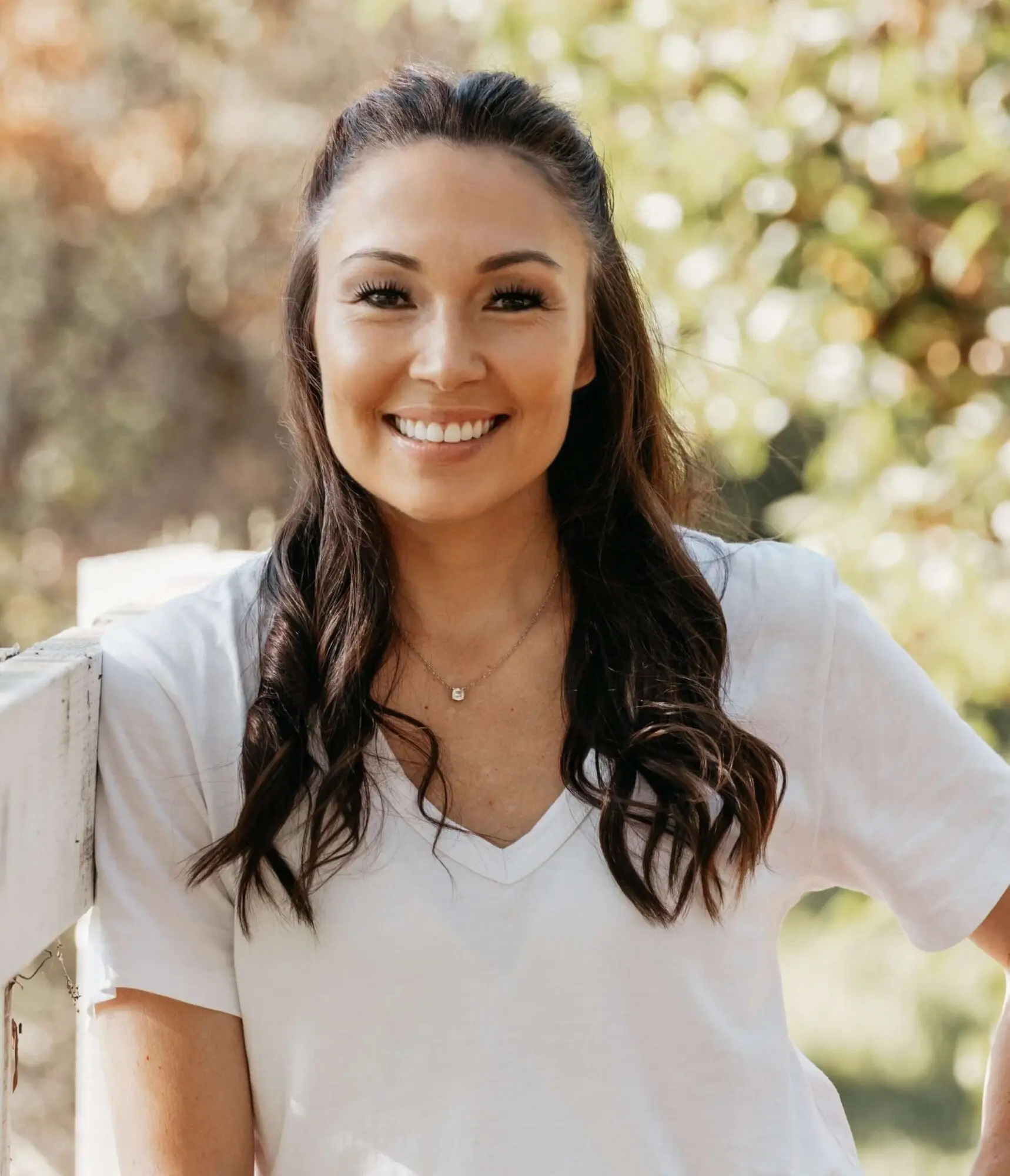At Stellar Orthodontics in San Marcos CA, Dr. Laura Deyo DDS, MS, a skilled orthodontist with long dark hair and wearing a white t-shirt, smiles outdoors by a white fence.