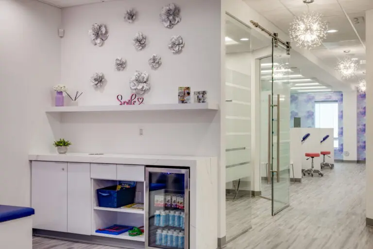 Stellar Orthodontics in San Marcos, CA features a modern, brightly lit office with white decor, wall art, glass doors, and a small fridge stocked with water bottles and supplies.