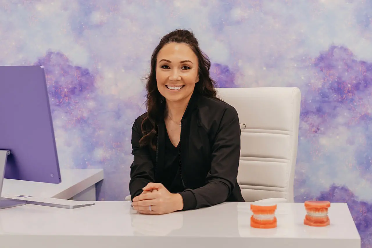 At Stellar Orthodontics in San Marcos CA, Dr. Laura Deyo, MS, an expert orthodontist, smiles beside dental models at her desk with a computer monitor, set against a purple patterned wall.
