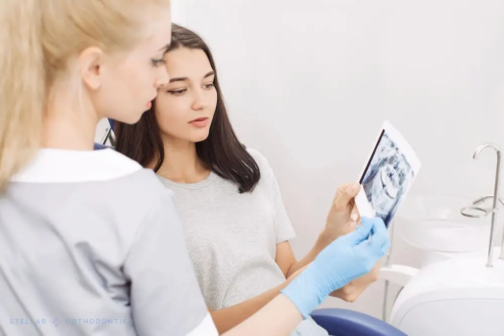 At Stellar Orthodontics in San Marcos, CA, a dentist in scrubs and gloves reviews a dental X-ray with a seated patient, discussing the results in the clinic after being explained about How to Fix Overbite in San Marcos, CA..