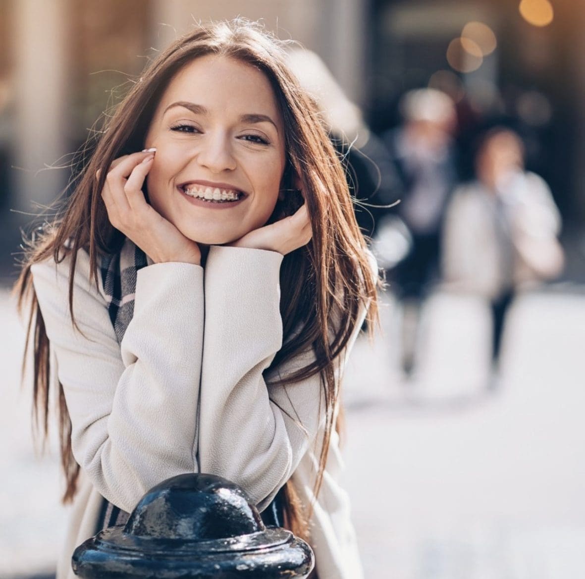 A young woman with long brown hair smiles confidently at the camera, resting her chin on her hands outdoors with blurred people in the background—highlighting the results of teen orthodontics at Stellar Orthodontics in San Marcos, CA.