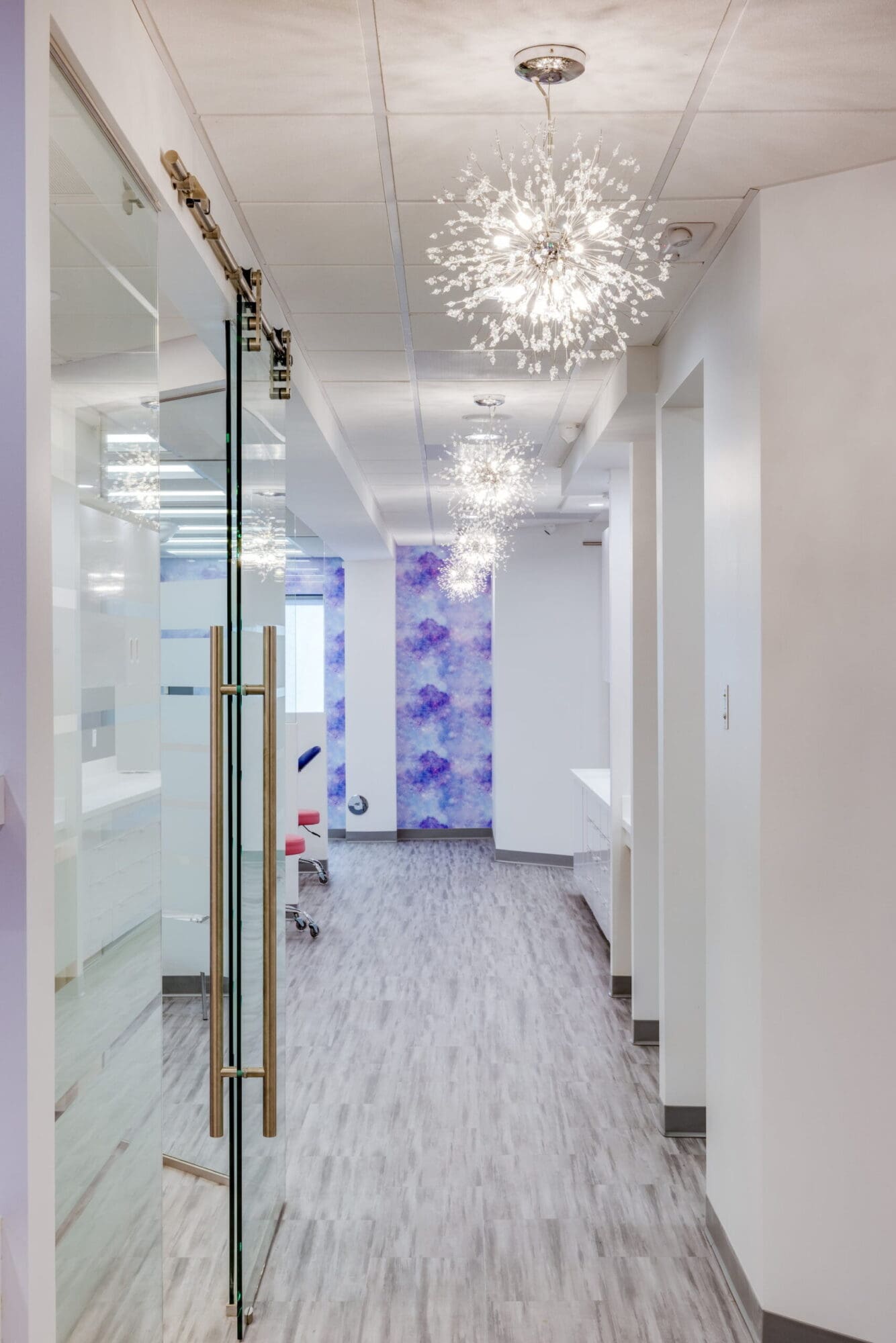 At Stellar Orthodontics in San Marcos CA, a modern hallway features glass doors, white walls, decorative ceiling lights, and purple abstract art at the end.