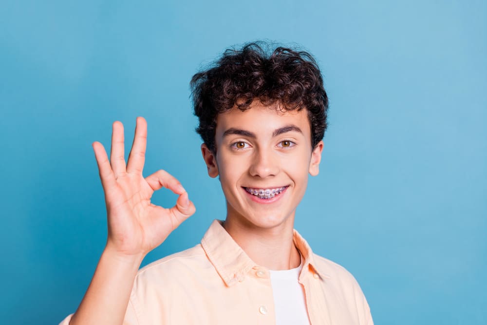 A teenage boy with braces flashes an "OK" hand sign against a plain blue background, representing Stellar Orthodontics in San Marcos, CA.