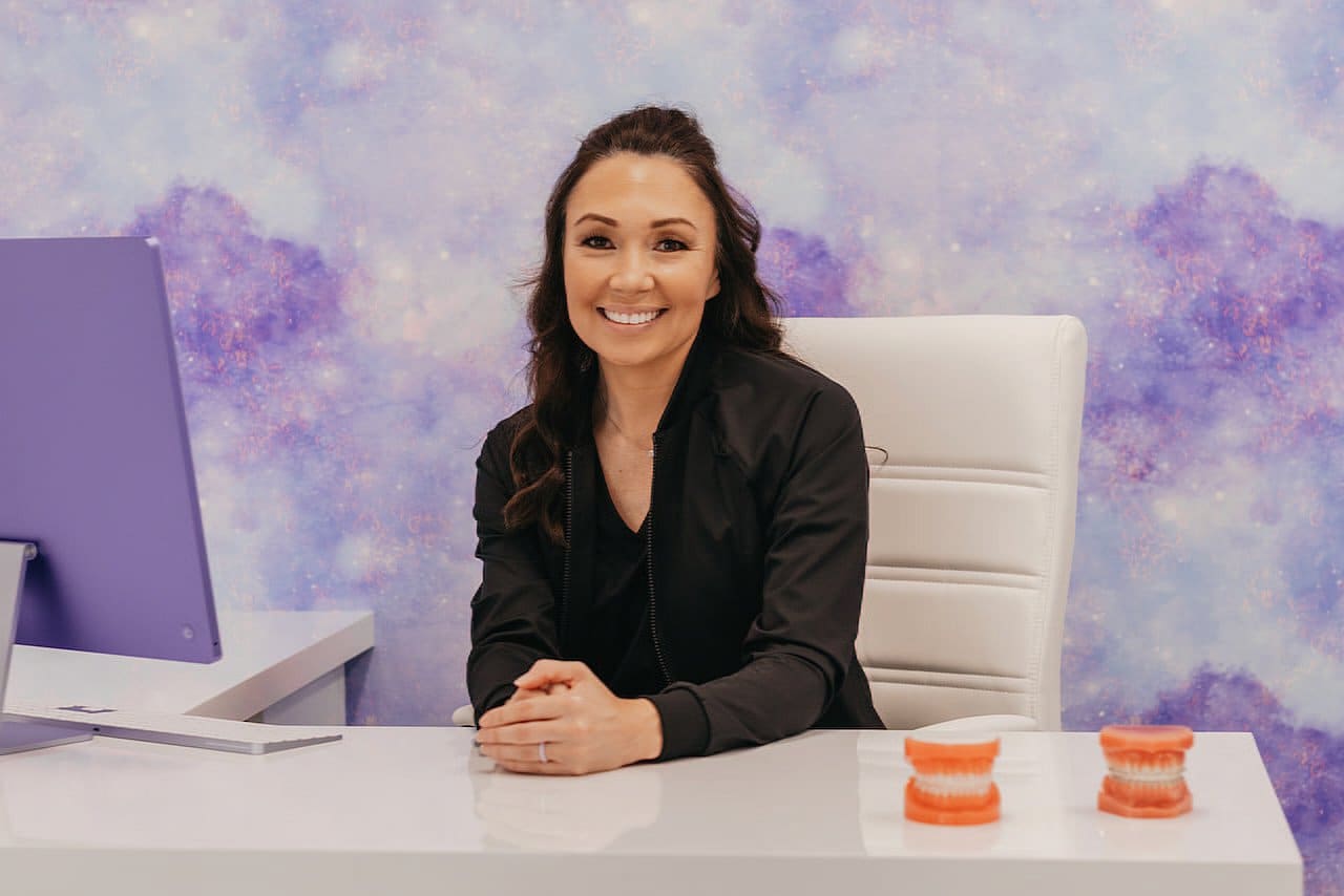 At Stellar Orthodontics in San Marcos CA, Dr. Laura Deyo, MS, an expert orthodontist, smiles beside dental models at her desk with a computer monitor, set against a purple patterned wall.