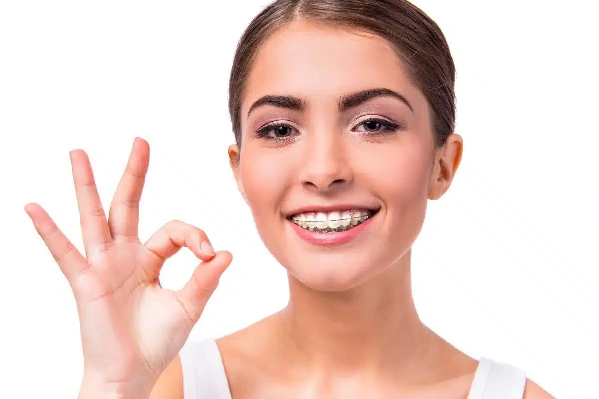 Smiling with braces, a young woman gives an "OK" gesture, promoting clear braces Stellar Orthodontics in San Marcos, CA, on white background.