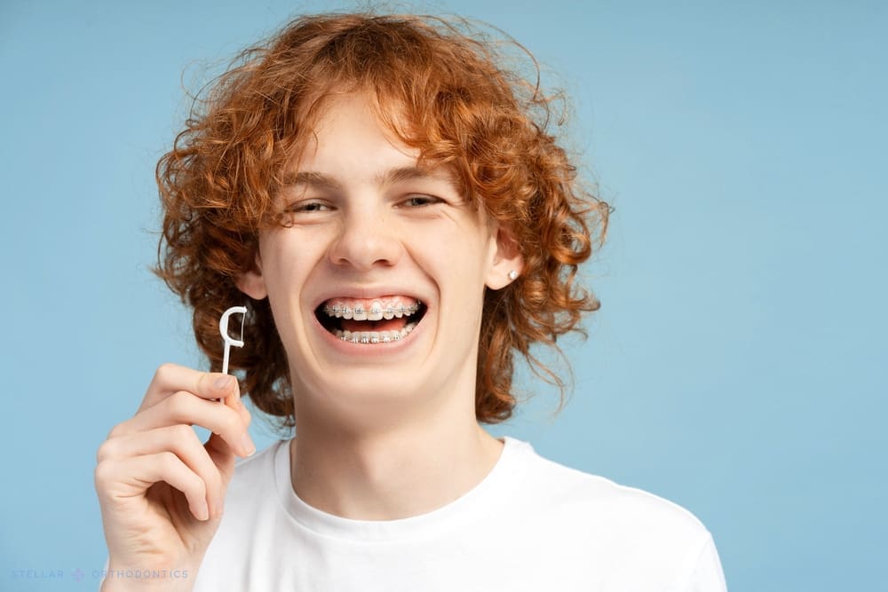 A smiling teen with curly hair shows metal braces and a dental flosser for Stellar Orthodontics in San Marcos, CA, on blue - How to Floss with Braces in San Marcos, CA