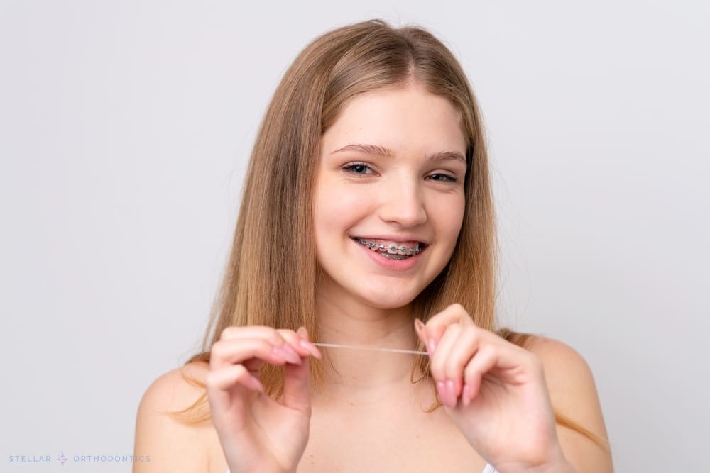 Teenage girl with braces holding dental floss, smiling, at Stellar Orthodontics in San Marcos, CA, against white background - How to Floss with Braces in San Marcos, CA