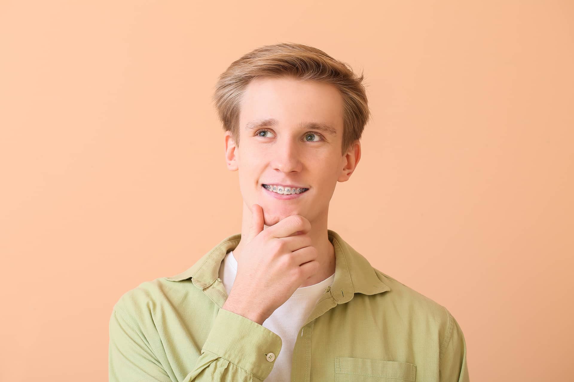 A young man with metal braces for Stellar Orthodontics in San Marcos, CA stands against a peach background, chin in hand, looking up.